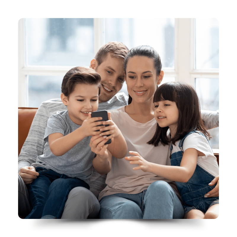 這張圖片展示了一個幸福的家庭在沙發上共同使用手機。父母與兩個孩子，男孩和女孩，圍坐在一起，專注地看著手機屏幕，顯示出家庭和睦和互動的場景。這說明了NAS在家庭中的應用，帶來了便捷的共享和存取數位內容的體驗。