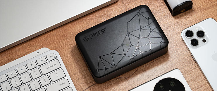 這張圖片展示了ORICO品牌的QIUDrive個人網路儲存器NAS設備，放置在木質桌面上。設備旁邊有鍵盤、筆記本電腦和手機，顯示出其在現代工作環境中的應用。QIUDrive以其便攜性和家用雲儲存功能聞名，提供方便的數據存儲和訪問解決方案，非常適合家庭和個人使用。