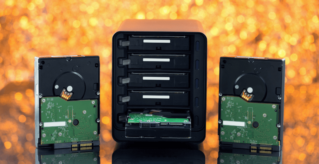 這張圖片展示了一個NAS設備的正面，設備內部可以看到插入的多個硬碟位。設備旁邊擺放著兩個取出的硬碟，顯示出NAS系統的可擴展性和易用性。背景為橙色模糊光點，強調了設備的技術感和現代感。這說明了NAS在數據存儲和管理中的重要性，提供了靈活的存儲解決方案。