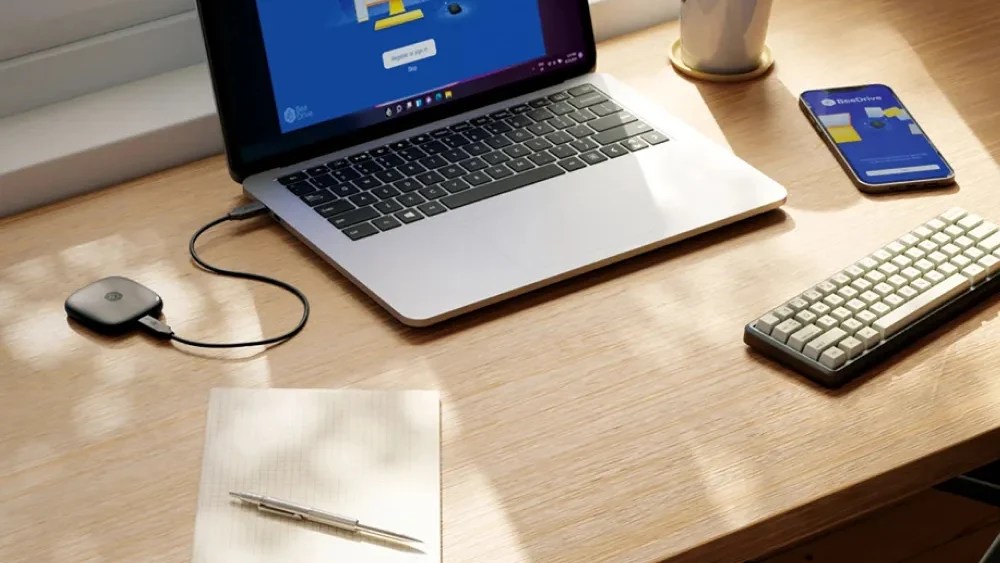 一台黑色的Synology 群暉科技 BeeDrive 2TB 個人行動備份裝置正在與一台輝似的型電腦傳輸資料，他們一同被擺放在靠在窗邊的木製書桌上